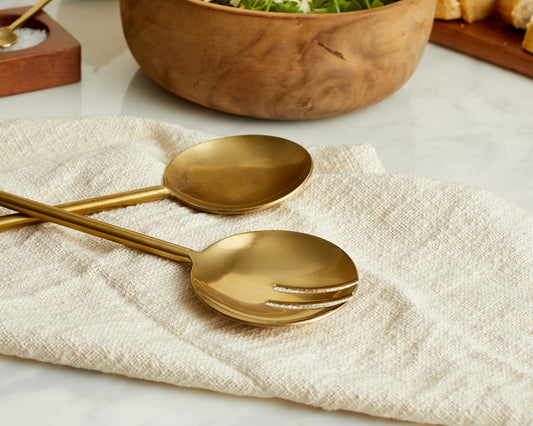Close-up of gold salad servers resting on a napkin beside a small salt dish and a salad bowl.