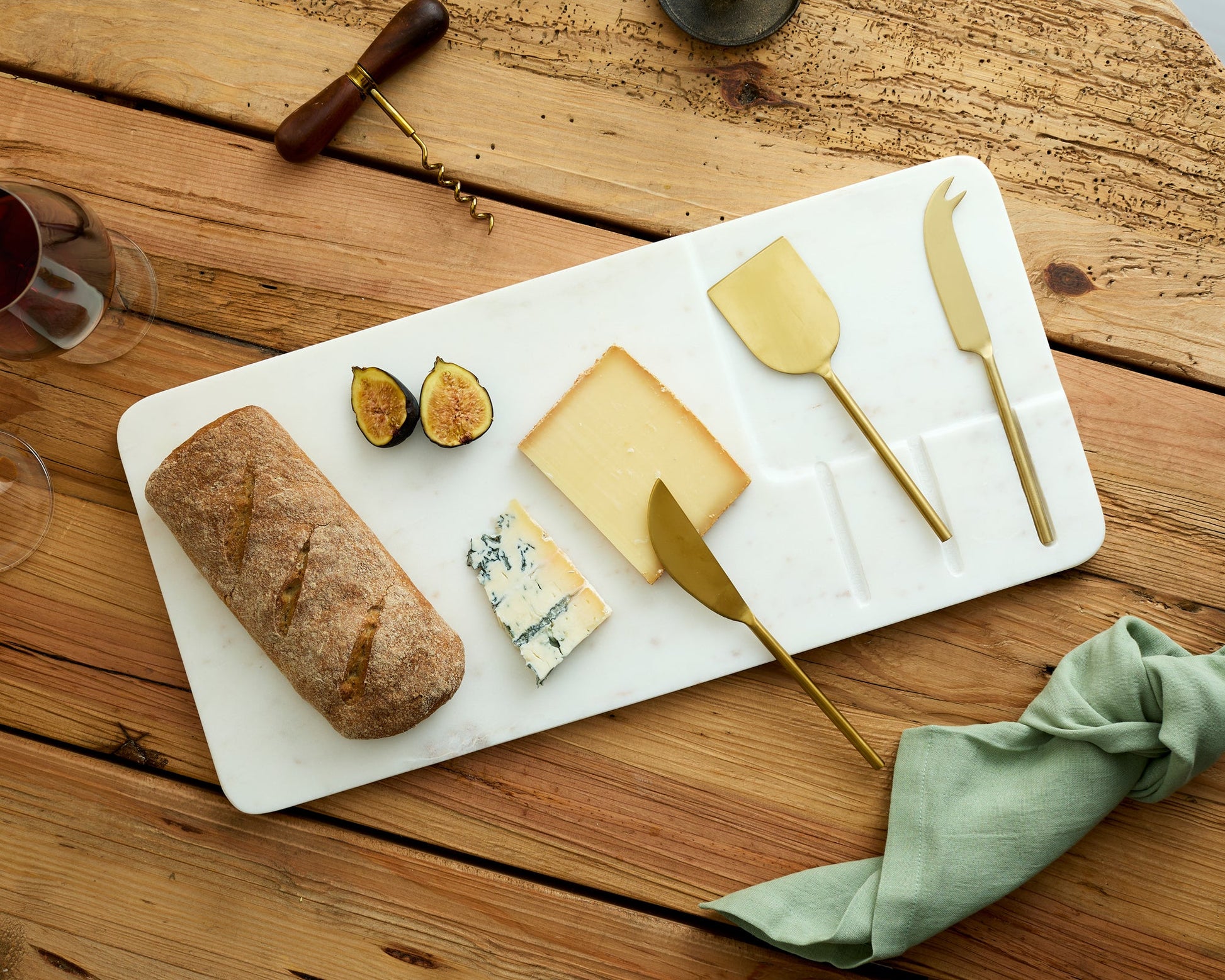 White marble cheese board with stainless steel cutlery set on wooden table with bread, fig and cheese