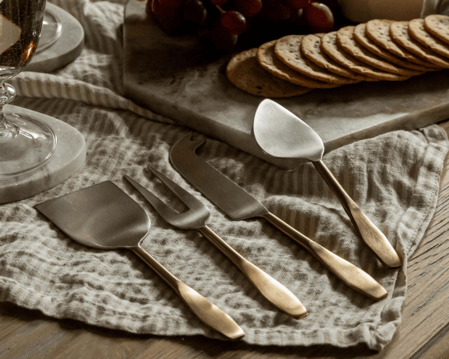 Edith cheese cutlery set 4 piece stainless steel serving set on linen tablecloth with cheese board and wine glass in background
