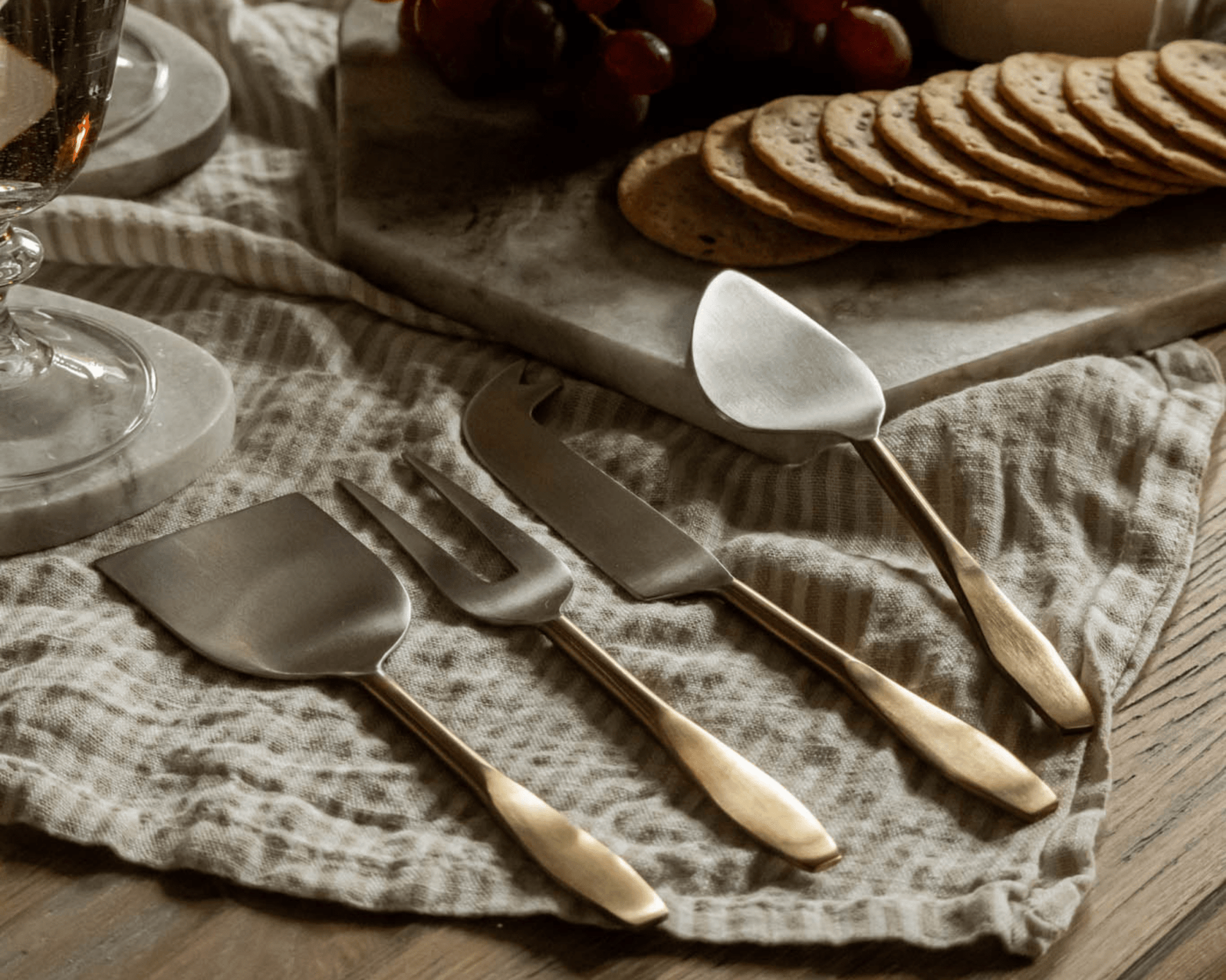 Edith cheese cutlery set 4 piece stainless steel serving set on linen tablecloth with cheese board and wine glass in background
