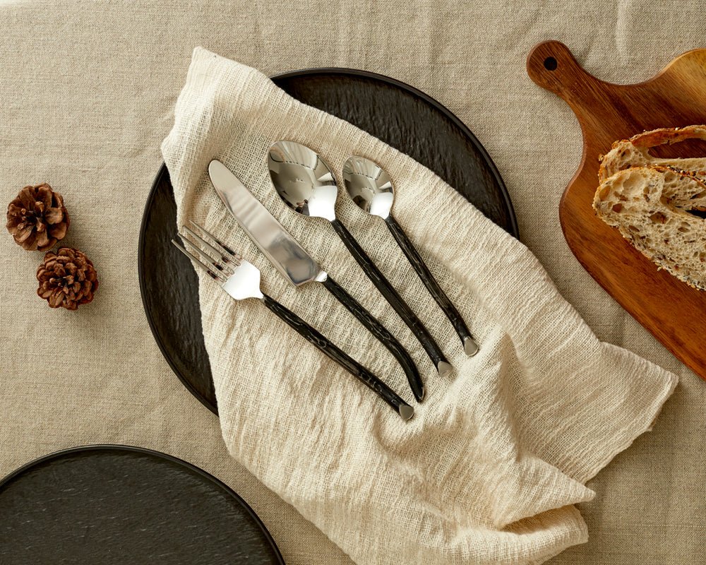 Silver cutlery set stainless steel Elmswell Rustic displayed on linen napkin on black plate with bread board to the side
