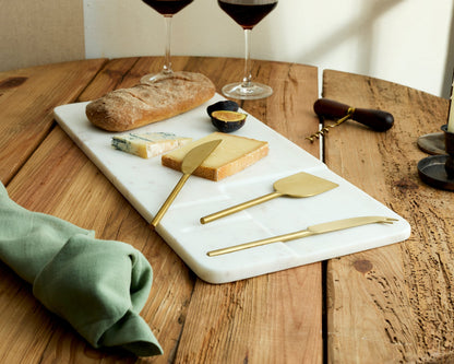 Angled view of white marble cheese board with stainless steel cutlery set, bread, fig, cheese, candles and wine glasses
