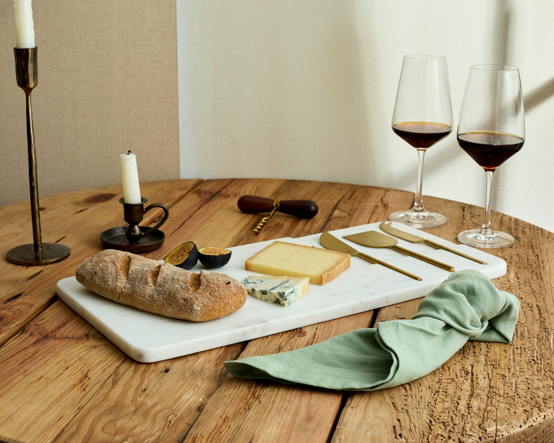 White marble cheese board with stainless steel cutlery set on wooden table with bread, fig, cheese, candles and wine glasses