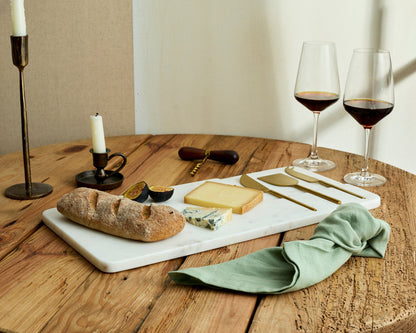 White marble cheese board with stainless steel cutlery set on wooden table with bread, fig, cheese, candles and wine glasses