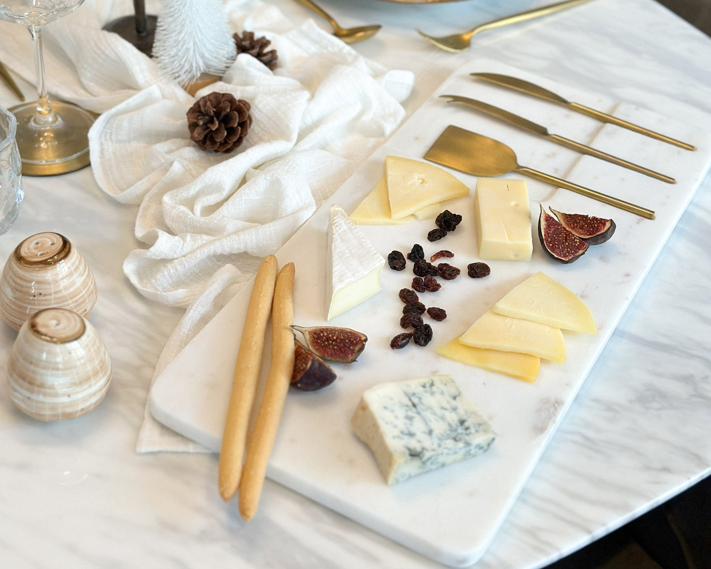 White marble cheese board with stainless steel cutlery set on wooden table with cheese, raisins, fig and breadsticks