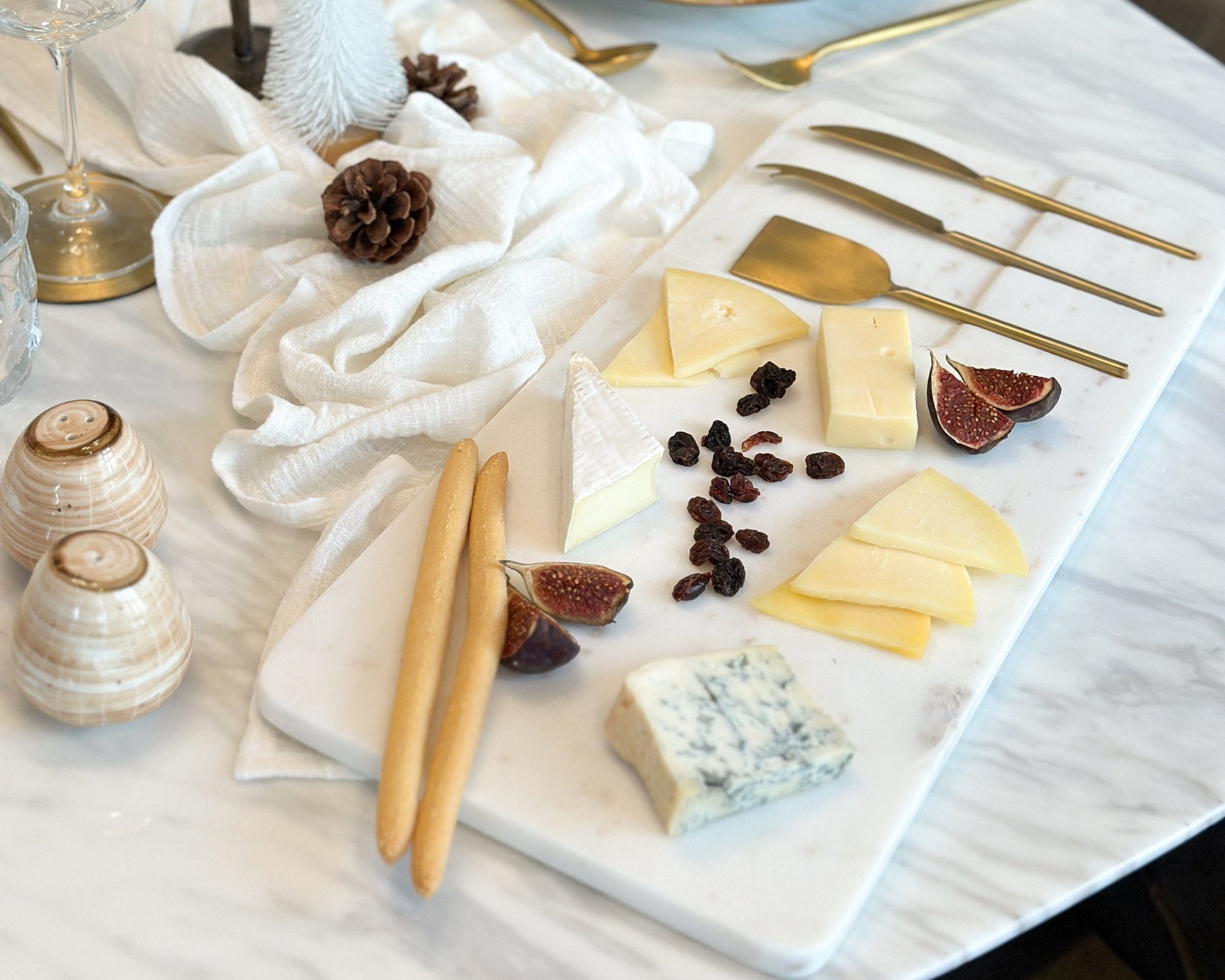 White marble cheese board with stainless steel cutlery set on wooden table with cheese, raisins, fig and breadsticks